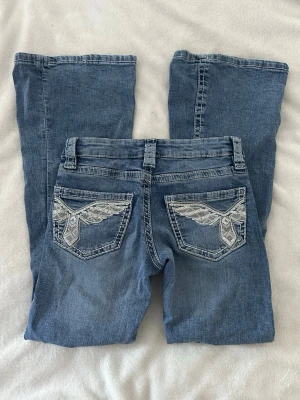 Blå jeans med broderade fickor Gina Tricot - Snygga slitna blå jeans från Gina Tricot med utsvängda ben och coola broderade detaljer på bakfickorna. Klassisk femficksmodell med bälteshällor och knapp med blommönster. De är i storlek 152 cm och är nästan aldrig använda.