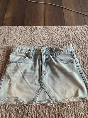 Ljus jeanskjol från Gina Tricot - Säljer en ljusblå jeanskjol från Gina Tricot i storlek 158. Kjolen har råa kanter, slitningar och klassiska fickor fram och bak. Den har en snygg omlott-look framtill och stängs med knapp och dragkedja.
