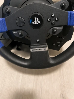 Thrustmaster ratt och pedaler PS4/PS5 - Säljer en Thrustmaster ratt och pedaler till PlayStation. Ratten har svart och blå design med PlayStation-knappar och detaljer i plast och syntetmaterial. Perfekt för racing och bilspel på PS4 och PS5. Kopplas enkelt in med USB.