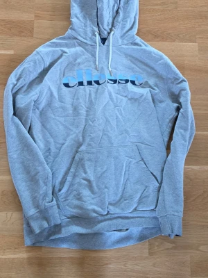 Grå hoodie från Ellesse med tryck - Snygg grå hoodie från Ellesse med stort blått och svart logotryck på bröstet. Klassisk huva med vita dragsnören och en stor magficka framtill. Tillverkad i mjukt bomullsmaterial och har ribbade muddar vid ärmar och nederkant.
