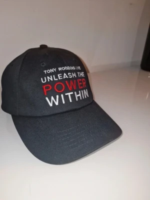 Svart keps Unleash The Power Within - Snygg svart keps med böjd skärm och broderad text fram: 'Tony Robbins Live Unleash The Power Within' i vitt och rött. På baksidan står det 'Breakthrough Success Resources' i blått och vitt. Justerbar rem bak för perfekt passform. Perfekt accessoar för dig som gillar personlig utveckling.