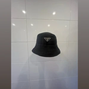Svart bucket hat från Prada - Snygg svart bucket. Hatten har en klassisk form och är tillverkad i ett glansigt, vattenavvisande material som ger en modern och clean look. Perfekt accessoar för att lyfta din outfit.