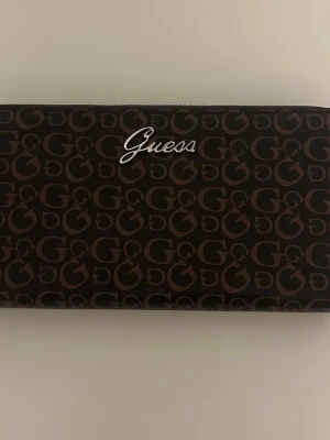 Brun plånbok med logomönster Guess - Snygg brun plånbok från Guess med all-over logomönster i mörkbrunt och Guess-logga i silver framtill. Plånboken har en rektangulär form och är tillverkad i ett tåligt syntetmaterial. Perfekt för dig som gillar stilrena accessoarer med tydlig branding.