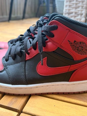 Svarta och röda Air Jordan 1 sneakers - Säljer ett par klassiska Air Jordan 1 sneakers i svart och rött skinn med ikonisk vinge-logga på sidan och röd swoosh. Skorna har högt skaft, svarta skosnören och vit mellansula. Perfekta för dig som gillar streetwear och vill sticka ut.