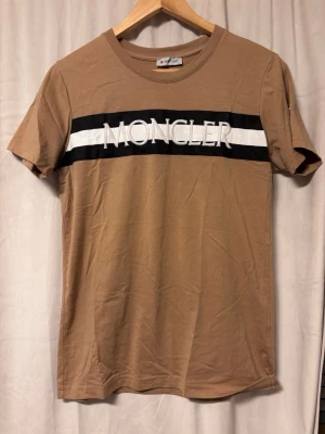 Moncler t-shirt - Snygg beige t-shirt  med vit och svart rand över bröstet och stor logga. Klassisk rund halsringning och korta ärmar. Märke även på ärmen. Tillverkad i mjuk bomull för skön känsla. Pris kan diskuteras vid snabb affär 