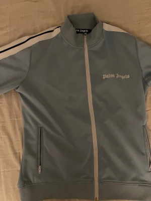 Ljusblå ziptröja från Palm Angels - Snygg ljusblå ziptröja från Palm Angels med vita och svarta ränder längs ärmarna. Tröjan har hög krage, dragkedja framtill och två fickor med dragkedja. Palm Angels-logga broderad på bröstet. Perfekt för en sportig och trendig look.