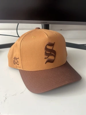 Brun och beige keps med S-brodyr - Snygg keps i beige med brun böjd skärm och stor S-brodyr framtill. På sidan finns en liten broderad detalj i brunt. Klassisk modell med justerbar baksida, perfekt för en streetwear-look. Tillverkad i slitstark bomull.