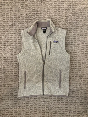 Grå fleeceväst från Patagonia - Snygg gråmelerad fleeceväst från Patagonia med hel dragkedja, hög krage och tre fickor med dragkedja. Västen har en stilren look och är perfekt för lager-på-lager. Materialet är mjukt och värmande, och detaljerna är i matchande grått.