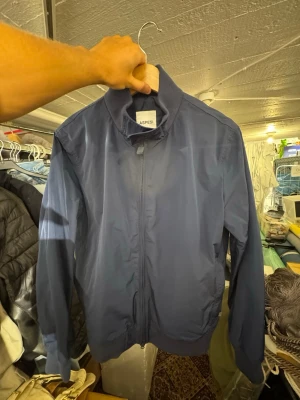 Marinblå vindjacka från Aspesi - Snygg marinblå vindjacka från Aspesi med dragkedja framtill och hög krage. Jackan har en minimalistisk design, ribbade muddar vid ärmslut och nederkant samt tillverkad i ett lätt, vindtätt material. Perfekt för blåsiga dagar. Finns att hämta i Gröndal. Nypris är 3049kr.