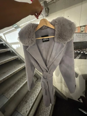 Ljusgrå kappa med fuskpäls Garoff - Snygg ljuslila kappa med bred krage i mjuk fuskpäls och knytskärp i midjan. Kappan har en klassisk siluett och är tillverkad i ett fluffigt material som ger en lyxig känsla. Perfekt för dig som vill sticka ut med en trendig och elegant look. Finns att hämta i Gröndal. 