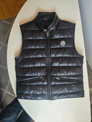 Svart dunväst från Moncler - Stilren orginal svart dunväst från Moncler med quiltad design och hög krage. Västen har två dragkedjefickor framtill och Moncler-logga på bröstet. Perfekt för lager-på-lager och håller dig varm med lätt dunfyllning.