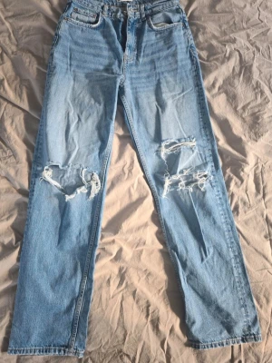 Ljusblå raka jeans med hål - Säljer ett par ljusblå jeans med raka ben och slitna detaljer över båda knäna. Jeansen har klassisk femficksmodell och hög midja. Perfekta för en avslappnad och trendig look.