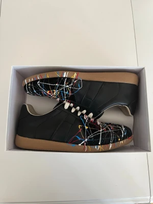 Svarta sneakers med färgstänk - Unika svarta sneakers med färgstänk i vitt, rött, blått och gult. Skorna har en klassisk låg siluett, vita skosnören och bruna gummisulor. Materialet ser ut att vara läder eller konstläder. Perfekta för dig som vill sticka ut med din stil.Pris (prutbart vid seriöst intresse).