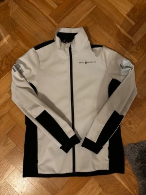 Vit och svart vindjacka Sail Racing XS - Snygg vindjacka från Sail Racing i vit och svart med hög krage och hel dragkedja framtill. Jackan har sportig design med svarta paneler längs sidor och ärmar, samt logga på bröstet och ryggen. Perfekt för dig som gillar stilren och funktionell look.