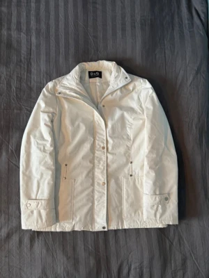 Vit Dolce&Gabbana jacka - Snygg vit vindjacka från Dolce Gabbana med hög krage och dragkedja samt tryckknappar framtill. Två fickor med dragkedja och justerbara muddar vid ärmsluten. Jackan har en clean och stilren look, perfekt för dig som gillar minimalistisk stil. Liten defekt med dragkedjan har gått av på ena fickan, men inget som märks. Skriv om frågor uppstår eller om t.ex. fler bilder efterfrågas.