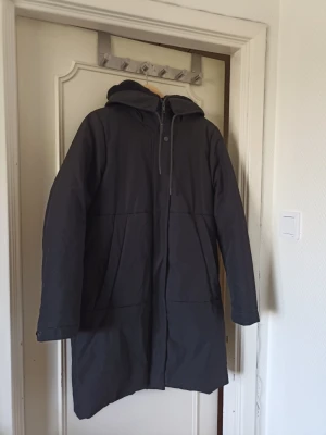 Mörkgrå lång parkas med huva - Snygg mörkgrå parkas med huva och dragkedja framtill. Jackan har två stora fickor på sidorna och en längre passform som går ner över låren. Perfekt för kalla dagar och enkel att matcha med olika outfits.