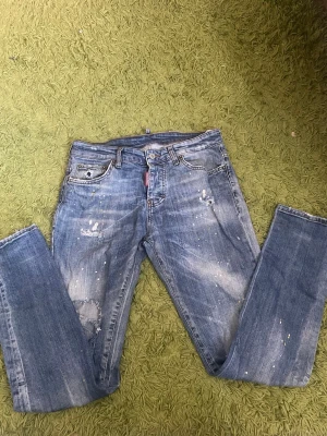 Blå Dsquared2 jeans med slitningar - Säljer ett par blå jeans från Dsquared2 med coola slitningar och färgstänk. Jeansen har raka ben, klassiska bakfickor och en tydlig Dsquared2-logga på ena fickan. 30/32. Perfekta för dig som gillar en streetig och avslappnad stil.