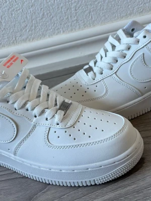 Nike Air Force 1 vita sneakers - Klassiska Nike Air Force 1 sneakers i helvitt läder med perforerad tå, vita skosnören och diskret Nike-logga på sidan och hälen. Tidlös design med robust sula och metallbricka på snörningen. Perfekta för en clean och stilren look.