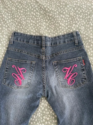 Blå jeans med rosa brodyr - Säljer ett par blå jeans med raka ben och coola rosa broderade initialer på bakfickorna. Jeansen har klassisk femficksdesign. Perfekta för dig som vill sticka ut med detaljer!