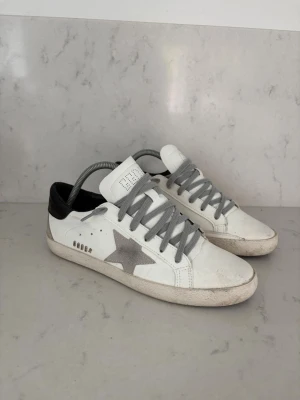 Golden goose  - Snygga vita sneakers med grå stjärndetalj på sidan, grå skosnören och svart hälparti. Skorna har en klassisk låg profil och är tillverkade i läder med gummisula. Perfekta för en avslappnad och trendig stil.