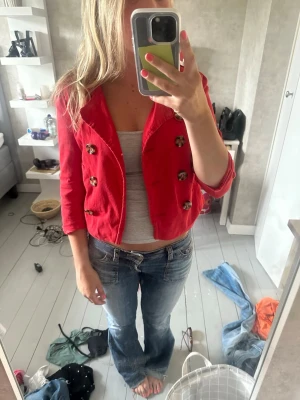 Röd kort kavaj med leopardknappar - Snygg röd kavaj i kort modell med unika leopardmönstrade knappar. Kavajen har trekvartsärmar och en figurnära passform som ger en trendig look. Perfekt att styla över ett linne eller t-shirt för en cool outfit.