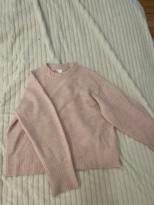 Ljusrosa stickad tröja - Mysig ljusrosa stickad tröja med rund halsringning och ribbade muddar vid ärmslut och nederkant. Tröjan har en avslappnad passform och är perfekt för kyliga dagar. Materialet ser ut att vara mjukt och behagligt.