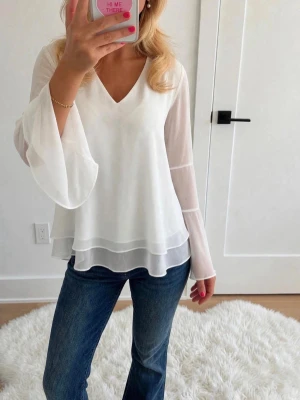 Vit v-ringad blus med vida ärmar - Säljer en superfin vit blus med v-ringning och långa, vida ärmar i transparent chiffong. Blusen har ett dubbelt lager tyg nedtill som ger ett flowy och luftigt intryck. Perfekt att styla med jeans för en trendig look.