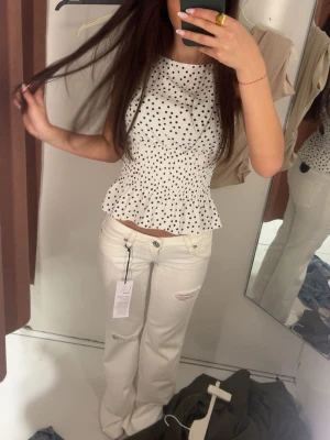 Vit prickig ärmlös topp med volang - Supersöt vit topp med svarta prickar och volangkant nedtill. Toppen är ärmlös och har en smockad midja som ger en snygg passform. Perfekt till jeans eller kjol för en trendig look.