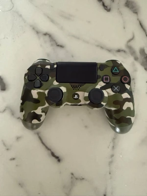 Kamo PlayStation-handkontroll - Snygg PlayStation-handkontroll med grön, beige och svart kamouflagemönster. Ergonomisk form med klassiska PlayStation-knappar och touchpad. Tillverkad i plast och passar perfekt för gaming-sessioner.