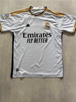 Real Madrid matchtröja vit/guld - En Real Madrid matchtröja från Adidas i vitt med svarta och guldiga detaljer. Tröjan har klubbmärke och sponsortryck på bröstet, samt La Liga-märke på ärmen. Tillverkad i lätt och ventilerande material, perfekt för fotbollsträning eller match.