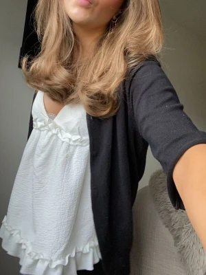 Vit volangblus med v-ringning - Söt vit blus med v-ringning och volangdetaljer längs bröstet och nederkanten. Blusen är ärmlös och har en lös passform som ger en avslappnad och trendig look. Perfekt att styla med en kofta eller jacka.