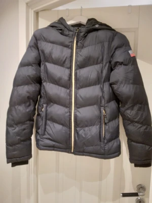 Svart pufferjacka / puffer med huva, strlk Small. Tjock och varm. - Snygg svart pufferjacka med huva och guldfärgade dragkedjor. Jackan har två fickor med dragkedja och en amerikansk flagga på ärmen. Quiltad design och ribbade muddar vid ärmsluten. Perfekt för kalla höst och vinterdagar. Finns även knappar på luvan så man kan köpa och sätta på pälskrage om man vill. Storlek Small. Fluffig och varm.