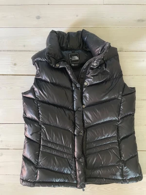 Svart dunväst från The North Face - Säljer en svart dunväst från The North Face med hög krage och tryckknappar framtill. Västen har ett glansigt material och klassisk quiltad design med snedställda sömmar. Perfekt för lager-på-lager och streetwear-stil. Gjord för kvinnor men funkar lika bra för män. 