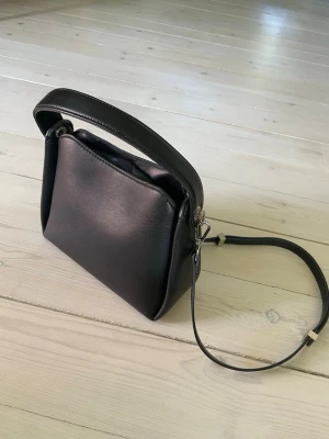 Svart handväska - Snygg svart handväska i skinnimitation med minimalistisk design. Väskan har både ett kort handtag och en avtagbar axelrem i samma material. Silverfärgade metalldetaljer och en strukturerad, boxig form ger ett modernt intryck.