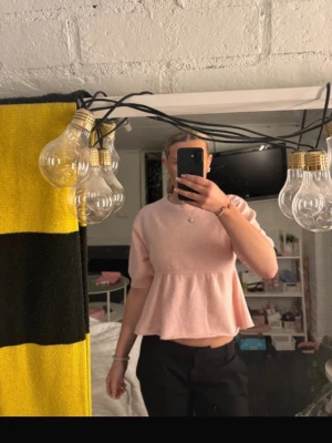 Rosa Nelly tröja - Söt ljusrosa topp med peplumvolang och korta ärmar. Toppen har en lös passform och är croppad, vilket ger en trendig och avslappnad look. Perfekt att styla med både jeans och kjol för en cool outfit.