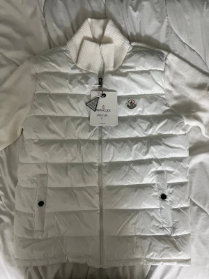 Vit dunväst från Moncler - Snygg vit dunväst från Moncler med ribbad krage och ärmar i stickat material. Västen har två fickor med knappstängning och dragkedja framtill. Klassisk Moncler-logga på bröstet och quiltad design för extra stilpoäng.