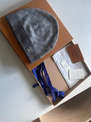Louis Vuitton mössa  - Snygg grå mössa från Louis Vuitton med diskret LV-logga framtill. Mössan är ribbstickad och har en uppvikt kant för extra stilpoäng. Perfekt accessoar för att hålla värmen och samtidigt se trendig ut.