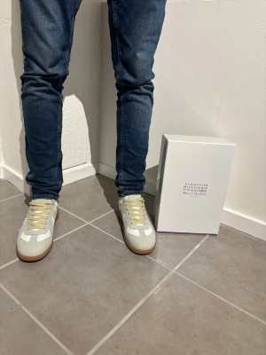Maison Margiela vita sneakers - Snygga vita sneakers från Maison Margiela med beige detaljer och gummisula. Skorna har en klassisk låg silhuett och är tillverkade i en mix av läder och mocka. Perfekta för dig som gillar stilrena och trendiga sneakers.
