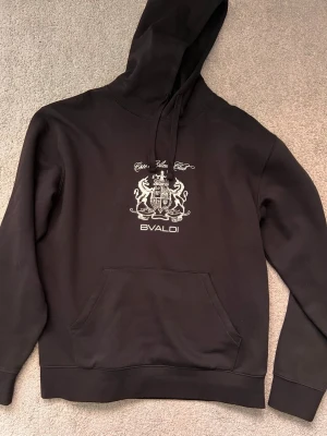 Svart hoodie från Bvaldi med tryck - Snygg svart hoodie från Bvaldi med vitt tryck på bröstet som visar ett stilrent emblem och texten 'Cote d'Azur Club'. Hoodien har huva med dragsko och en stor magficka. Perfekt för dig som gillar streetwear och vill ha en clean look.
