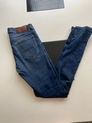 Mörkblå slim fit jeans herr - Snygga mörkblå slim fit jeans med klassiska fem fickor och kontrastsömmar. Jeansen har en läderpatch bak i midjan och subtila slitningar framtill för en trendig look. Tillverkade i denim med stretch för extra komfort.