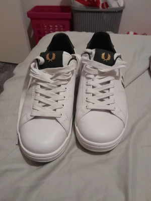 Vita Fred Perry sneakers med logga - Snygga vita sneakers från Fred Perry med klassisk svart logga på sidan och gulddetaljer på plösen. Skorna har vita snören, svart hälparti och är tillverkade i slätt läder. Perfekta för en clean och stilren look. Storlek 41
