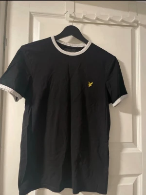 Svart t-shirt från Lyle & Scott - Snygg svart t-shirt från Lyle & Scott med vita kontrastkanter vid hals och ärmslut. Klassisk passform och gul logga på bröstet. Perfekt till jeans eller shorts för en clean och enkel stil.