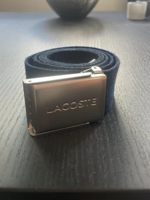 Ett flätat bälte från Jack and Jones och ett marinblått från Lacoste - Säljer två stilrena flätade bälten, ett i marinblått med en liten broderad detalj och ett i beige med bruna skinn-detaljer. Perfekta för att lyfta din outfit och passar till jeans eller chinos.