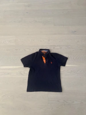 Mörkblå piké från Polo Ralph Lauren - Snygg mörkblå pikétröja från Polo Ralph Lauren med orange detaljer och klassisk krage. Insidan av kragen har ett rutigt mönster i blått, vitt och orange. Ikonisk logga broderad på bröstet. Perfekt för en avslappnad men stilren look.