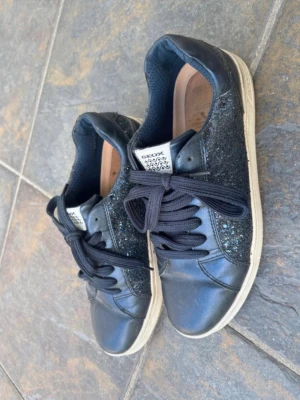 Svarta glittriga sneakers från Geox - Snygga svarta sneakers från Geox med glittriga sidopaneler och svarta skosnören. Skorna har vit sula och är tillverkade i en kombination av läder och textil. Perfekta för dig som vill ha lite extra bling i vardagen.