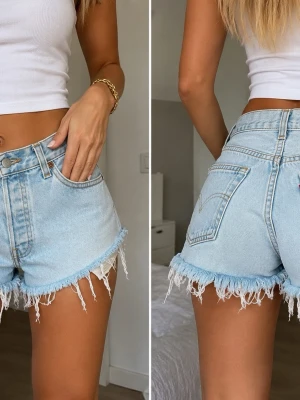 Levi’s 501 Original Jeansshorts – Ljus tvätt – S/M - Ljusblå Levi's 501 jeansshorts med hög midja och råa fransiga benslut. Klassisk femficksmodell i bomull med knappgylf och avslappnad loose fit. Perfekta för sommardagar och ger en chill vintagekänsla.