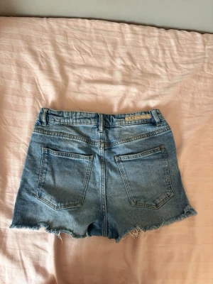 Blå jeansshorts med fransig kant - Säljer ett par klassiska blå jeansshorts med fransig, rå kant nertill. Shortsen har fem fickor, bälteshällor och knappgylf. Perfekta för varma dagar och ger en avslappnad vibe till din outfit.