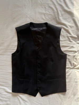 DRESSMANN SUPER 100 Premium kostymväst, S100 - Stilren svart kostymväst från Dressmann i super 100's ull. Västen har klassisk V-ringning, fyra knappar framtill och två fickor. Perfekt att matcha med kostymbyxor för en clean och dressad look. Original pris: 999kr
