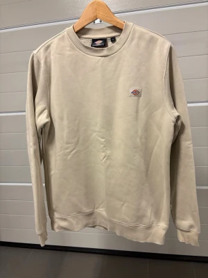Beige sweatshirt från Dickies - Snygg beige sweatshirt från Dickies med rund halsringning och diskret logga på bröstet. Tröjan har långa ärmar och ribbade muddar vid ärmslut och nederkant. Perfekt för en avslappnad och stilren look.