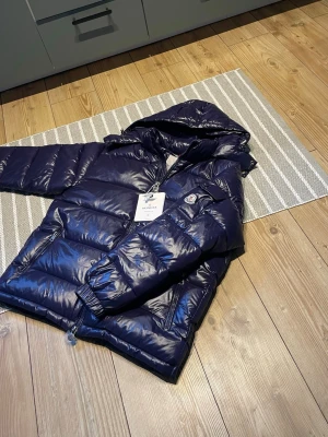 Mörkblå dunjacka från Moncler - Snygg mörkblå dunjacka från Moncler med glansig finish och huva. Jackan har två sidofickor med dragkedja, elastiska muddar vid ärmsluten och klassisk Moncler-logga på ärmen. Perfekt för kalla vinterdagar och riktigt trendig look.